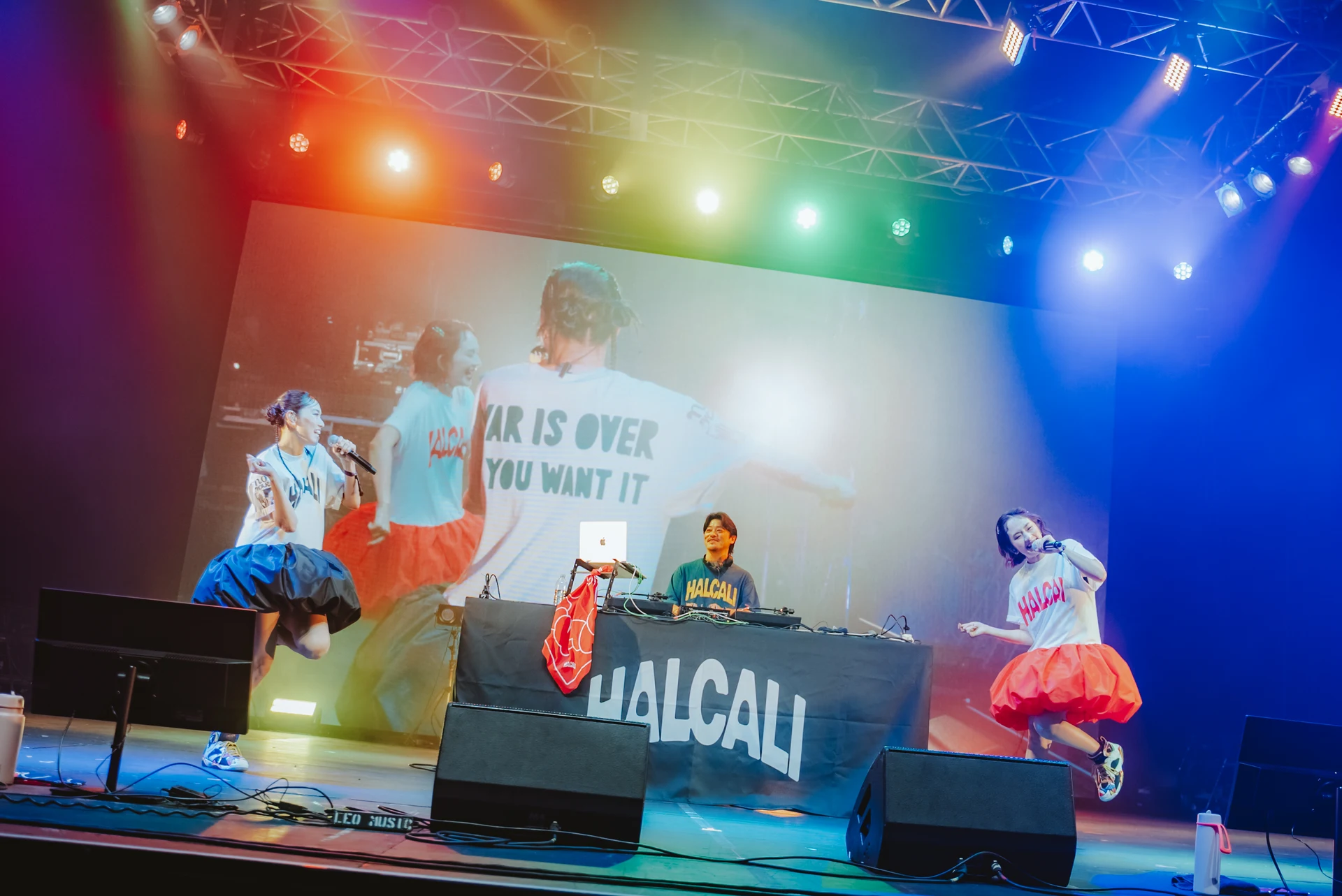 虹色照明のライブステージで、HALCALIの2人がマイクで歌い、中央のDJが演奏。背面スクリーンにWAR IS OVER YOU WANT ITと映る。