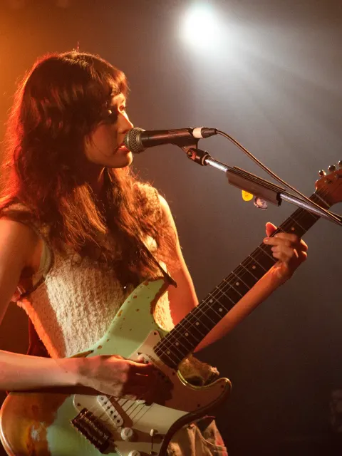A singer under a spotlight playing a pale green electric guitar and singing into a microphone on a stage lit in red and orange.