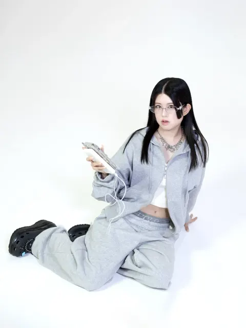 A woman with black hair and glasses kneeling in a gray outfit in a white studio, holding a wired smartphone with a cool expression.