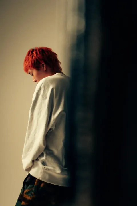 A red-haired person in a white sweatshirt standing in profile with their head lowered, against a pale interior wall and a dark, blurred foreground.