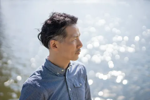 A man in a blue shirt stands in profile by the water, quietly looking into the distance. Sunlight sparkles on the softly blurred water behind him.