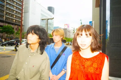 Three young people stand together in a city: a dark-haired man on the left, a blond man in a blue shirt in the center, and a woman dressed in red on the right. Tall buildings and road signs are in the background, with strong light washing out the right side.
