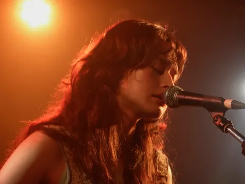 A long-haired singer performing into a microphone on an orange-lit stage.