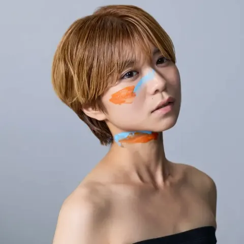 A person with short blond hair poses with bare shoulders against a blue background, wearing blue and orange paint on their cheek and neck and a calm expression.