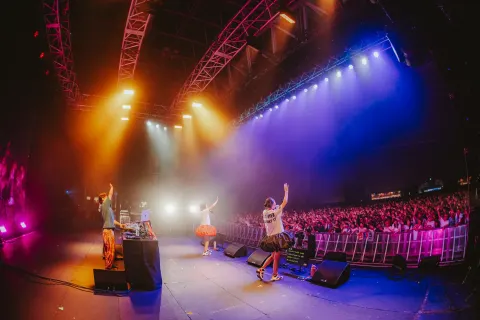 Three performers raising their hands onstage as a large audience cheers beneath purple and orange lighting.
