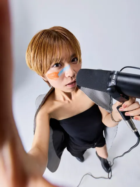 A selfie-like close-up of a woman with orange and blue face paint against a white background, looking at a black microphone held close to her face. The microphone is marked SHURE SM7B.