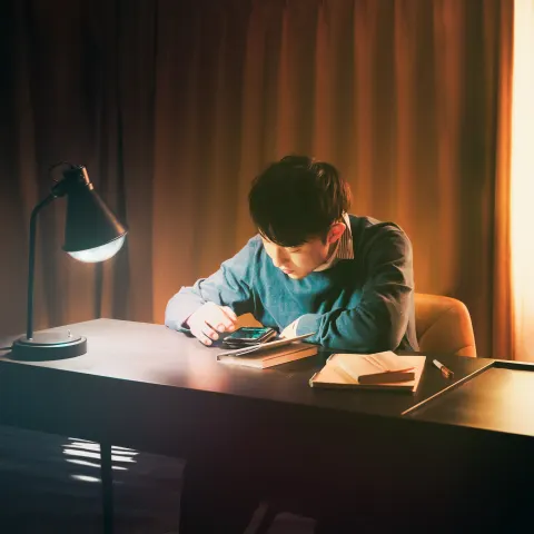 A young man studies while looking at his phone at a warmly lit desk. A black desk lamp, notebook, and curtains create a quiet, focused atmosphere.