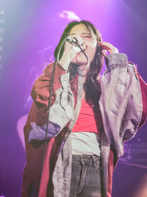 A singer in red and gray singing passionately into a handheld microphone with one hand against her ear in a purple-lit venue.