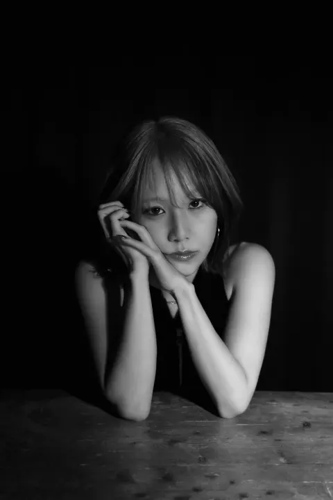 A black-and-white portrait of a woman resting a hand against her cheek in front of a wooden table and black background, with a quiet, pensive expression.