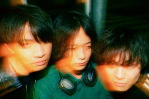 Three young men with headphones around their necks lean their faces close together, wearing serious expressions in dim green and orange light.