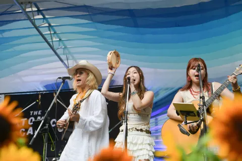 A man in a hat and two women sing and play on an outdoor stage under a blue canopy. The person in the center raises a tambourine, and the person on the right plays guitar. A sign reading “THE…” stands at the back left.