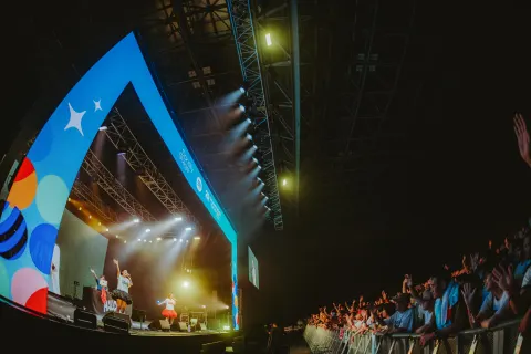 Singers and a DJ performing beneath a huge blue stage arch in an indoor venue, with audience hands raised, bright lighting and HALC lettering onstage.