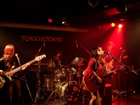 A four-piece of vocals, guitar, bass and drums performing in front of a TOKIO TOKYO sign in a red-lit live house.