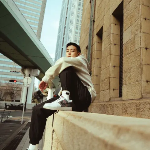 On an urban sidewalk beneath an elevated structure, a man in a beige hoodie and black trousers sits on a stone edge, with white sneakers visible.