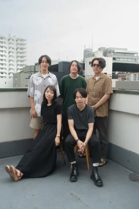 Five people posing on a rooftop, some sitting and some standing with calm expressions against a backdrop of city buildings.