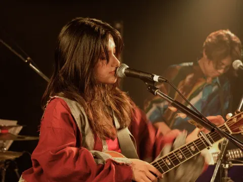 A singer in red playing guitar and singing on a warmly lit stage, with another musician performing behind her.