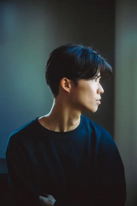 A dark-haired man in black clothing, lit by blue light, looks outside in profile from indoors, with a quiet, contemplative expression.