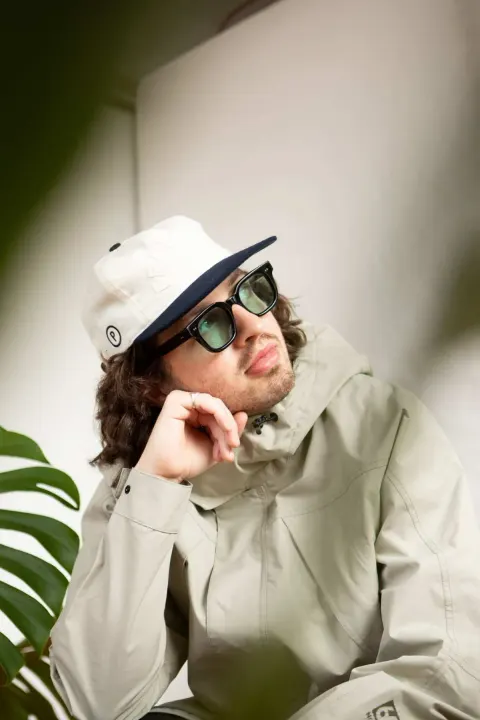 Indoors, a man in a white cap with a P logo, black-framed sunglasses, and a light beige jacket rests his chin on his hand and quietly looks upward past a plant.